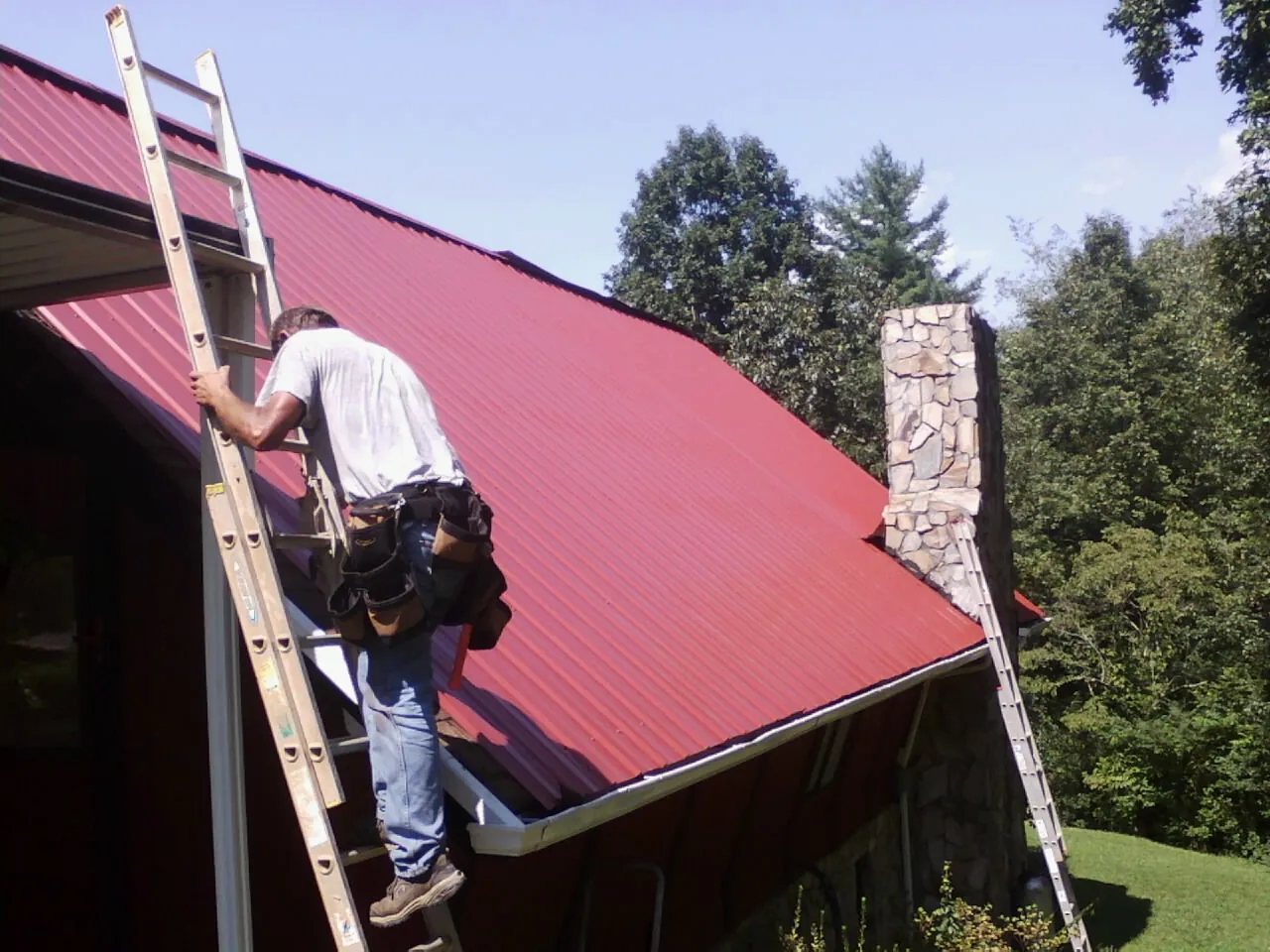 Roofer on a ladder beside a red metal roof and stone chimney — Soffit & Fascia Repair by Roofing Brothers in Saline